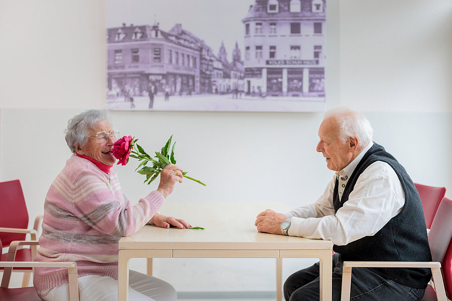 Zwei ältere Menschen sitzen sich an einem Tisch gegenüber, eine Frau hält eine Blume vor ihr Gesicht