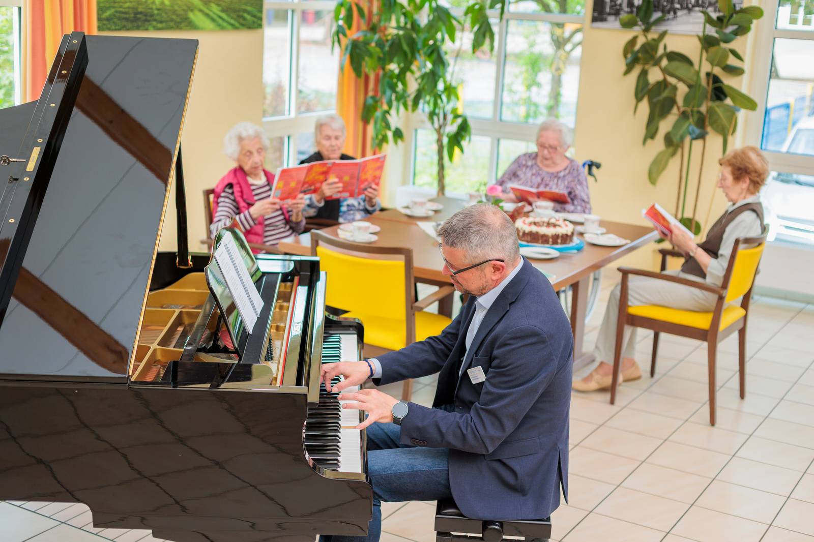 Pianist spielt am Klavier in einem hellen Raum, im Hintergrund ältere Personen mit Büchern und Kuchen.